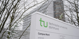 Ein mit Schnee bedecktes Schild mit TU-Logo und im Hintergrund steht das Mathematikgebäude.