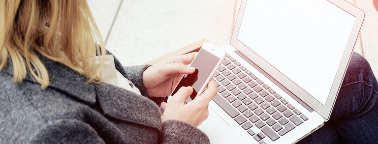 Frau mit Laptop auf dem Schoß, hält ein Smartphone in den Händen.