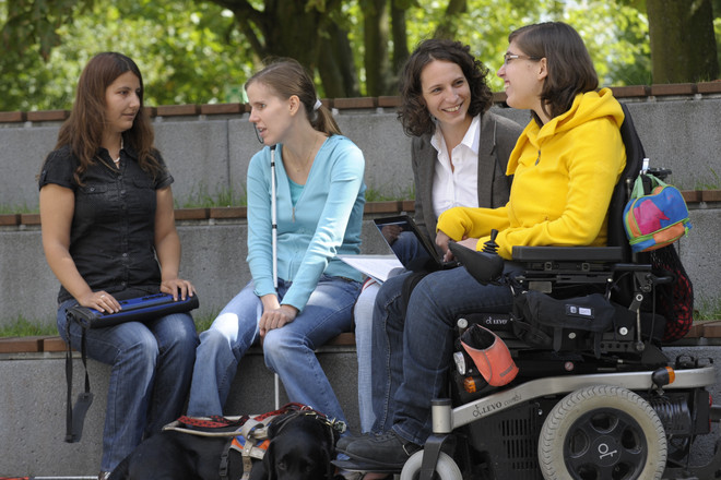 Vier Frauen sitzen auf einer Bank auf dem Campus und unterhalten sich. Zwei von ihnen haben eine körperliche Beeinträchtigung.