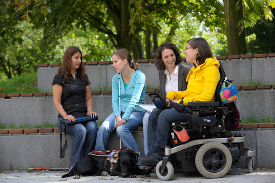 Vier Frauen sitzen auf einer Bank auf dem Campus und unterhalten sich. Zwei von ihnen haben eine körperliche Beeinträchtigung.
